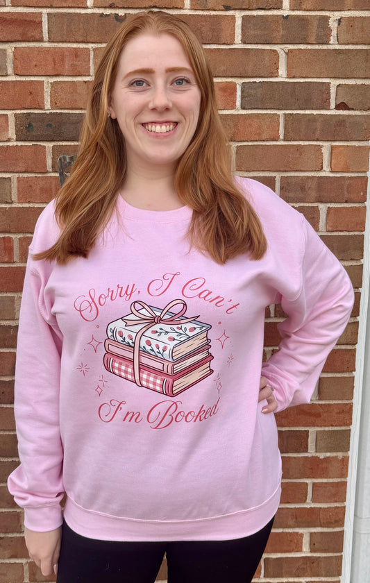 Person wearing a pink sweatshirt with a book-themed design and text, standing against a brick wall.