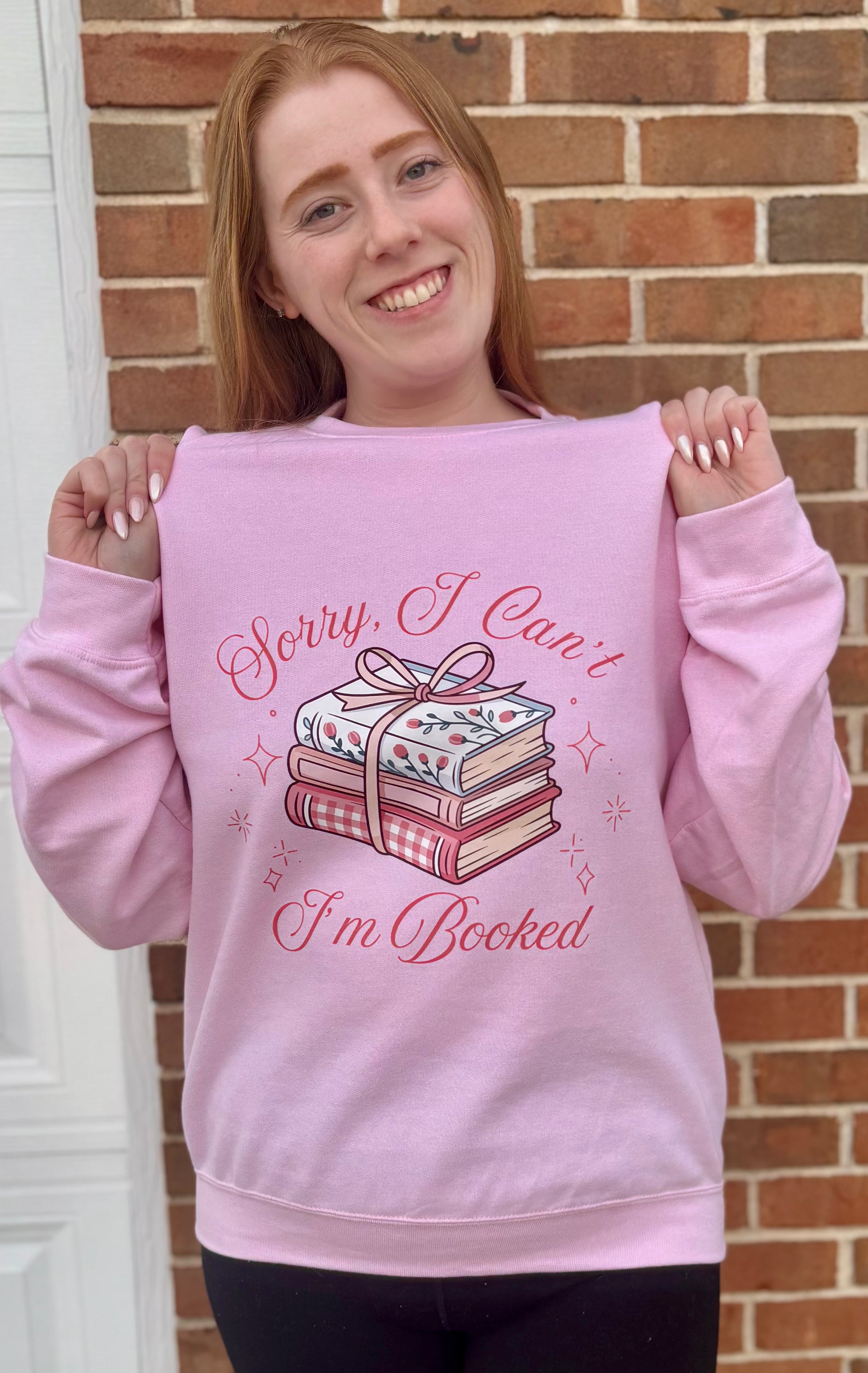 Person wearing a pink sweatshirt with a book-themed design in front of a brick wall.
