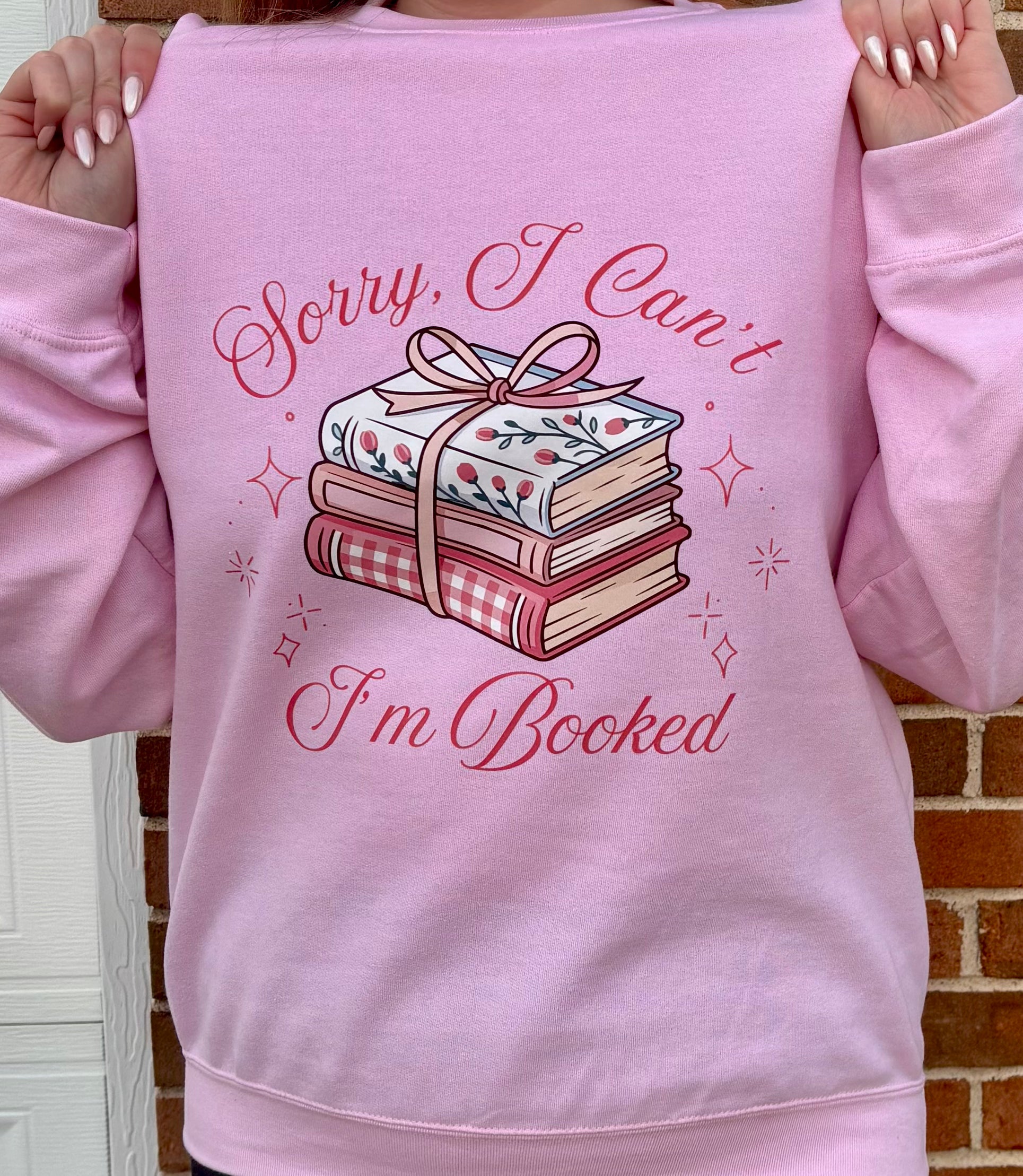 Pink sweatshirt with book-themed design and text held by a person against a brick wall.