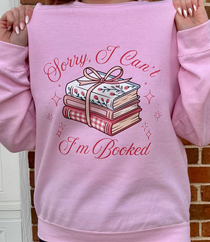 Pink sweatshirt with book-themed design and text held by a person against a brick wall.
