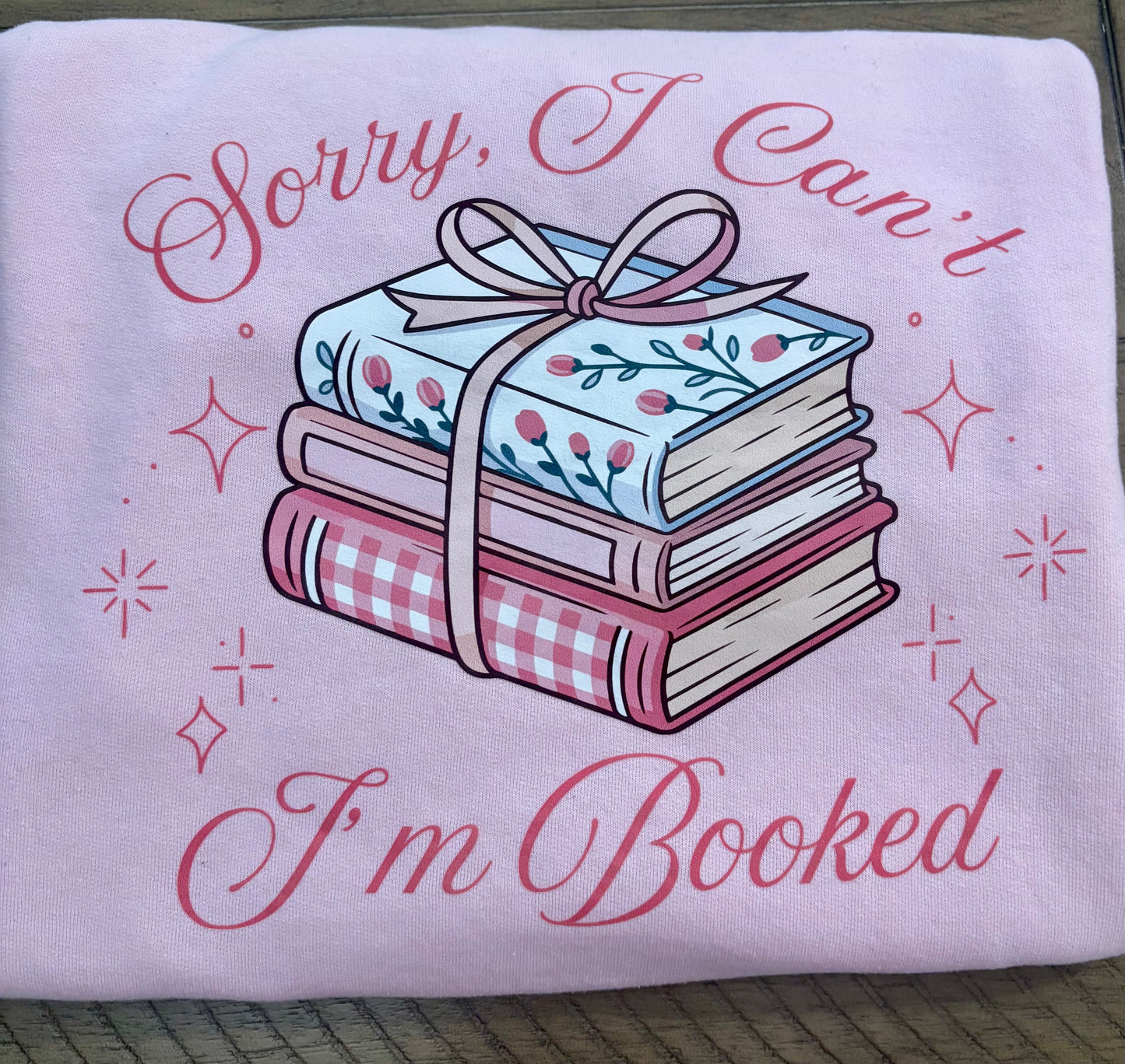 Pink folded sweatshirt with books and a bow design and text 'Sorry, I Can't I'm Booked' on a wooden surface.