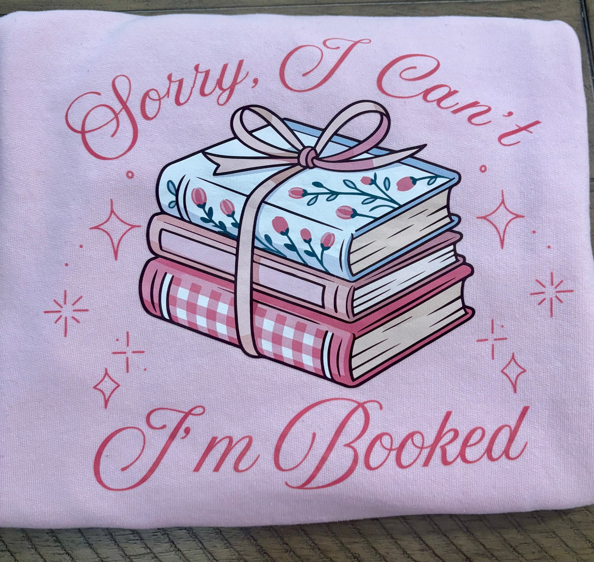 Pink folded sweatshirt with books and a bow design and text 'Sorry, I Can't I'm Booked' on a wooden surface.