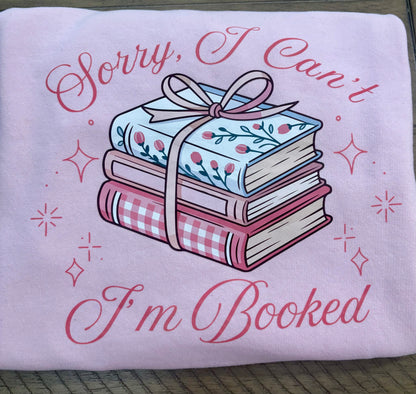 Pink folded sweatshirt with books and a bow design and text 'Sorry, I Can't I'm Booked' on a wooden surface.